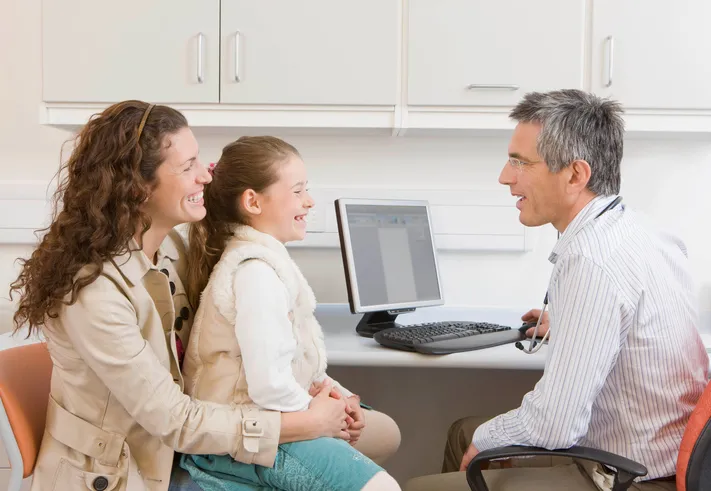 daughter sitting on mother's lap talking with doctor at his desk